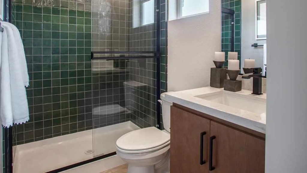 A modern bathroom with green tiled walls, a glass shower, wooden vanity, and white towels.