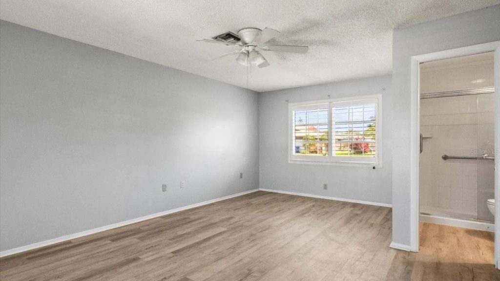 A calm bedroom with light gray walls, light wood floors, a large window with white shutters, a ceiling fan with lights, and an attached bathroom with a glass shower.