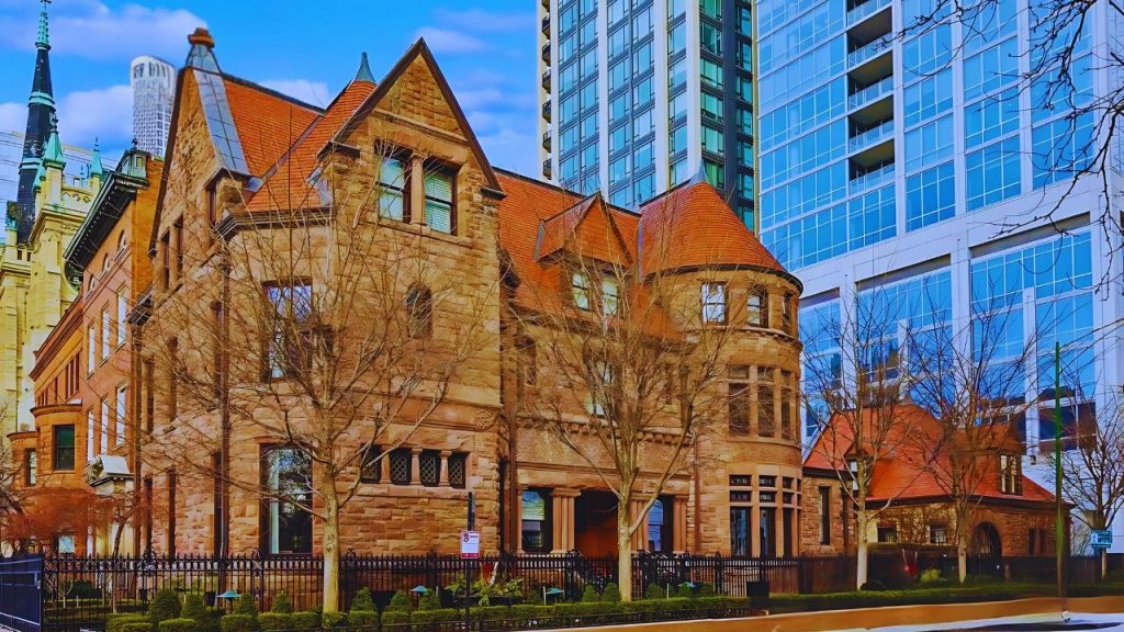 Grand stone mansion with red roof in downtown Chicago, surrounded by modern high-rises.