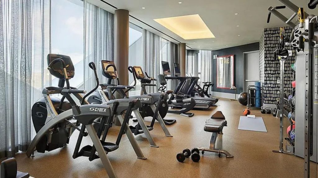 A modern gym with Cybex machines, weights, and large windows offering a city view.