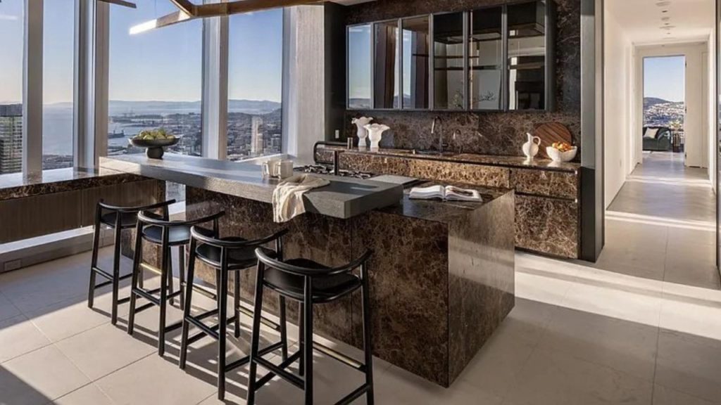 A modern kitchen with a marble island, black stools, and large windows showcasing a city skyline view.