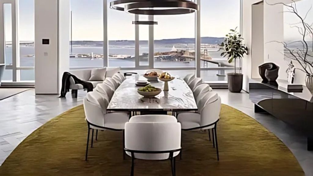 A stylish dining area with a marble table, white chairs, and large windows overlooking a bay view.