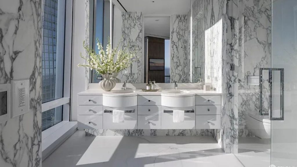 A luxurious bathroom with marble walls, a double vanity, and large windows offering a city view.