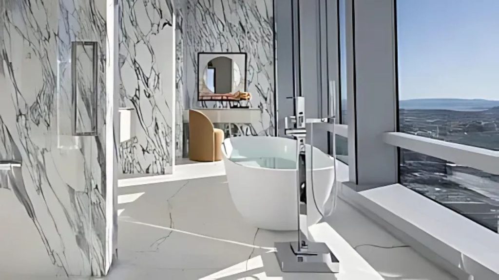 A serene bathroom with a marble tub, large windows, and a city view, featuring a cozy chair and mirror.