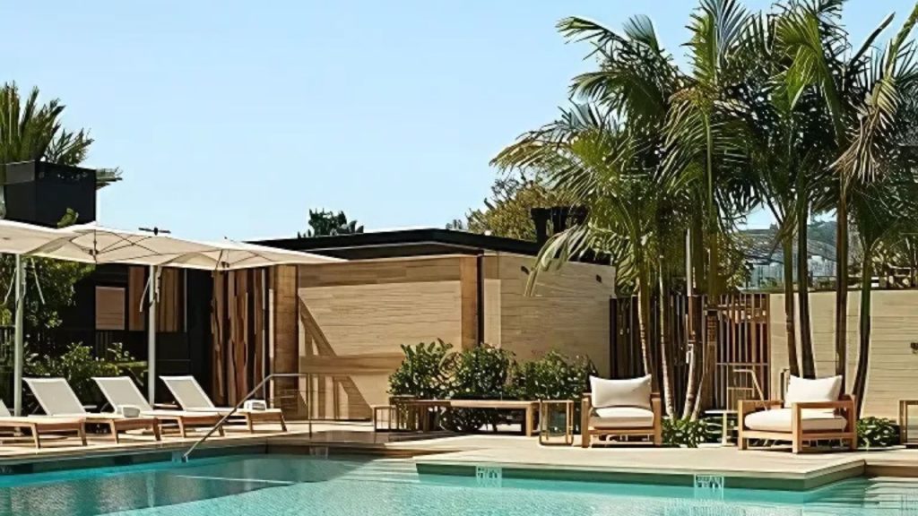 A tropical poolside with lounge chairs, a shaded cabana, palm trees, and clear blue water.