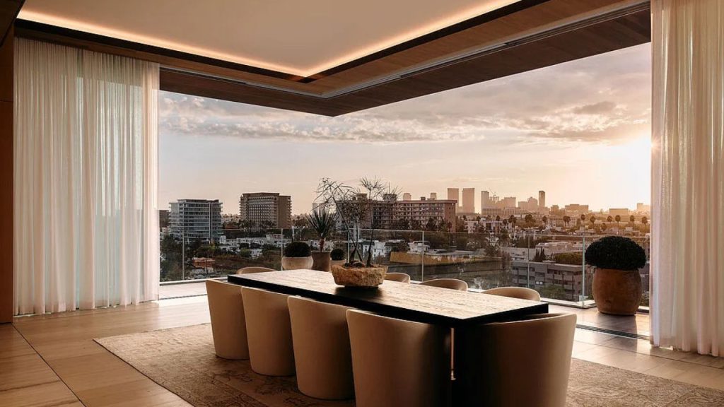 A sophisticated dining area with a long table, beige chairs, and a city skyline view at sunset.