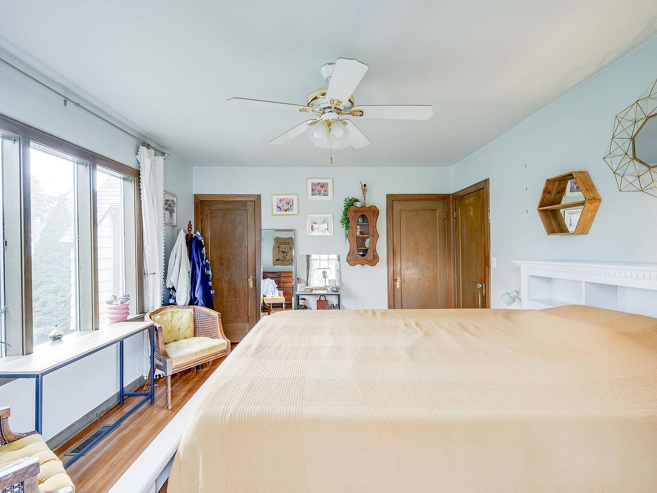 Bedroom with beige bedspread, wooden doors, and large windows with white curtains